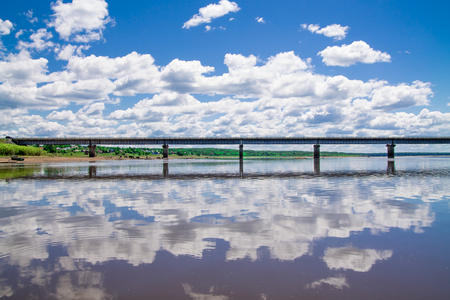 steel bridge with lots of fluffy clouds in the bright blue sky and their reflection on still water like in a mirrorの写真素材