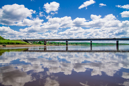 steel bridge with lots of fluffy clouds in the bright blue sky and their reflection on still water like in a mirrorの写真素材