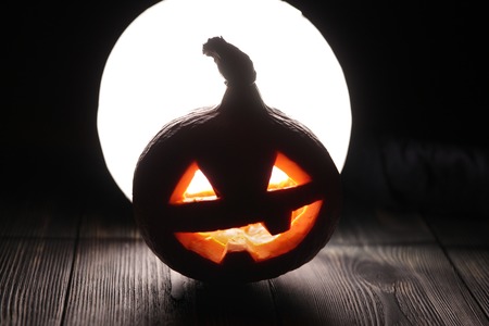 Halloween pumpkin Jack-o-lantern on the dark wooden background with a scary face as a symbol of halloween night with the concept full moon behind it.の写真素材