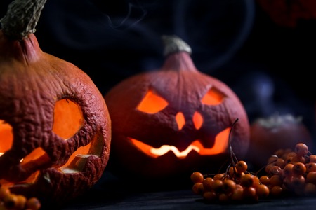 scary halloween pumpkin lanterns with luminous scary faces with rowanberries on a wooden background and the clouds of smoke in front of themの写真素材