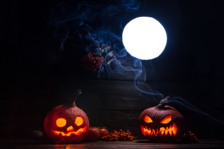 mystical Halloween luminous pumpkin Jack-o-lanterns with scary faces, a full moon with the clouds of smoke and rowanberries bunches on the dark wooden backgroundの写真素材