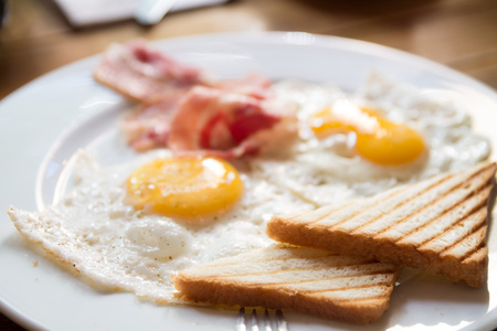 breakfast with fried eggs, bacon, toasts and cappuccino in white dishes on a wooden tableの写真素材