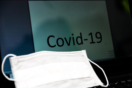 word Covid-19 written on LED monitor of a notebook with aprotective medical mask on keyboard