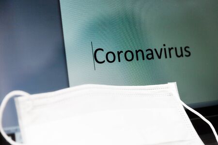 word Coronavirus written on LED monitor of a notebook with a protective medical mask on a keyboardの写真素材
