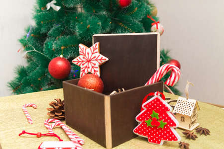 Dark brown box with gingerbread and sugar cane candies inside and Christmas toys next to it on golden surface, with Xmas fur tree on background. Happy New Year 2021の写真素材