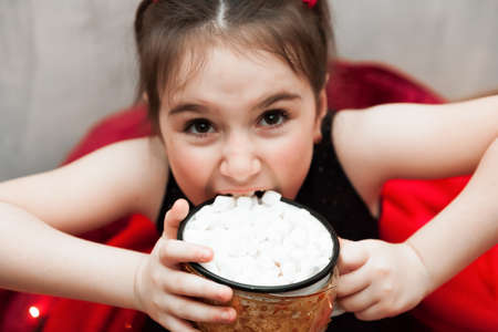 Defocused Funny little Girl Holding Cup with Hot Tasty Chocolate and Marshmellow and trying to eat it on cozy Red Christmas Blanket on Christmas Eve. Flat Lay Top View Stilllifeの写真素材
