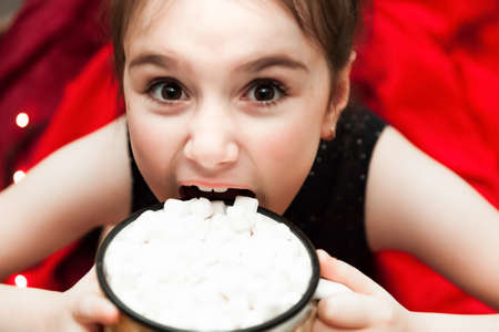 Defocused Funny little Girl Holding Cup with Hot Tasty Chocolate and Marshmellow and trying to eat it on cozy Red Christmas Blanket on Christmas Eve. Flat Lay Top View Stilllifeの写真素材