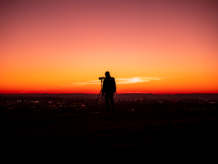Image of a silhouette of a man with tripot and camera standing on a hill during colorful sunsetの写真素材