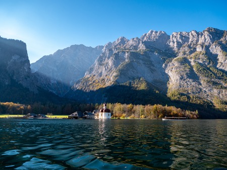 Image of the Koenigssee with the chapel of st bartholomew and a tourist boat. In the background the illuminated mountain formation of Watzmann in bavaria, Germanyの写真素材