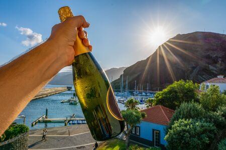 One hand holding a bottle of champagne in front of a marina during sunsetの写真素材