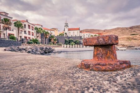 OLd rusty bollard on a stone pier with blurry village in the backgroundの写真素材