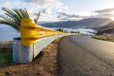Yellow guardrail along curvy road with sunset over the sea and villageの写真素材