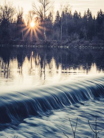 Water flowing over a weir during sunset with sun rays in the winter, Augsburgの写真素材