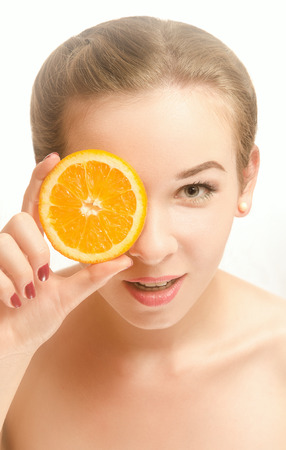 Portrait of an attractive young woman with a slice of orange in front of her eyes, which stands on white background and smilingの写真素材