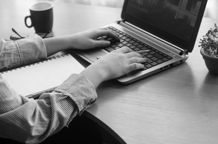 Young woman's hands working at a laptop, black and white image. Cup of coffee, notepad, pen,...