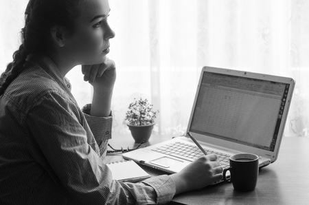 Young woman lost in thought, sitting at a laptop, black and white, pencil in hand, beside lying notebook, flowerpot and cupの写真素材