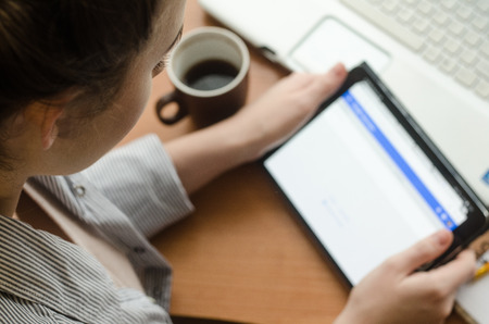 Young woman sitting at a laptop and holding a a tablet in hands. Nearby stands a coffee cupの写真素材