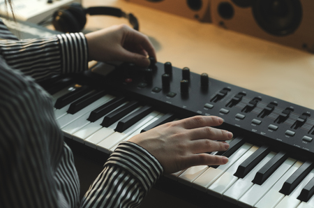 A woman in a striped shirt plays the synthesizer and tunes it. The body in the frame. Nearby are headphones, a music console and speakers. Side viewの写真素材