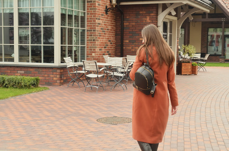 The girl in a coat and backpack on her shoulder, with long hair, goes to the coffee table, back to the photographer. Around beautiful buildingsの写真素材