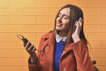 A young European woman standing against a background of a brick wall and listening to music on headphones with the phone.  Dressed in a sweater and a coat. The background is orangeの写真素材