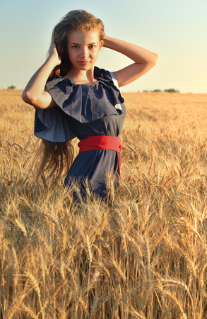 A beautiful young woman with long hair standing in the wheat field. Touches her hair, looks at the camera. Vertical photoの写真素材