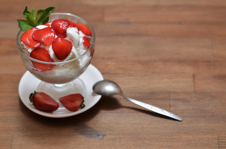 Vanilla ice cream with sliced strawberries and mint in a glass bowl on a white saucer with a teaspoon and halves of strawberries. Stand on a dark wooden tableの写真素材