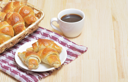 Croissants on a plate and in a basket, coffee in a cup, on a light wooden table with a napkinの写真素材
