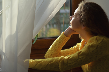 Portrait of a beautiful girl who looks out the window sitting in a yellow sweater at home, horizontal photoの写真素材