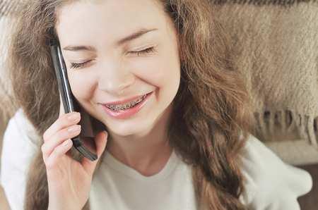 Cheerful brunette girl talking on mobile phone while sitting on comfortable couch. Smiling hipster girl having cell telephone conversation while resting near sofaの写真素材