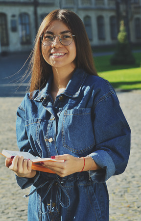 A student in a denim suit stands on the territory of the University and holds an open book in her hands, looks smiling at the cameraの写真素材