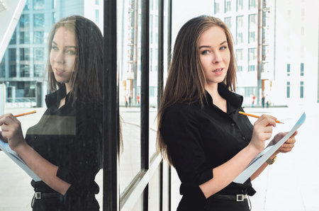 A beautiful young girl reflects on a complex technical project, standing outdoor near the building, writing down ideas on paperの写真素材