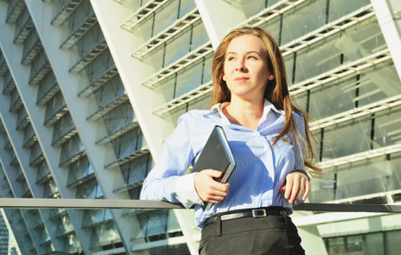 Young beautiful business lady standing in office clothes leaning on the railing, and holding a...