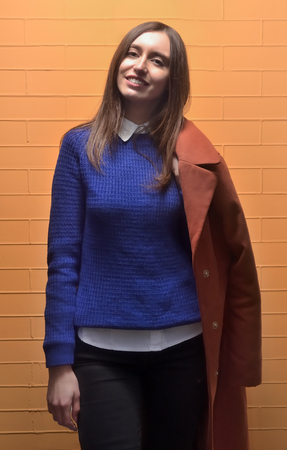 Portrait of a young pretty girl who is wearing a blue sweater with a white shirt, with a coat that hangs on her shoulder. The girl smiles and looks at the camera, standing near a brick wall of orange colorの写真素材