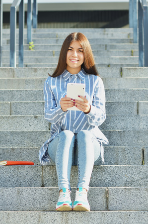 A young girl of Asian appearance, a student dressed in jeans and sneakers, holding a tablet and sitting on the stairs, and communicates by videoの写真素材