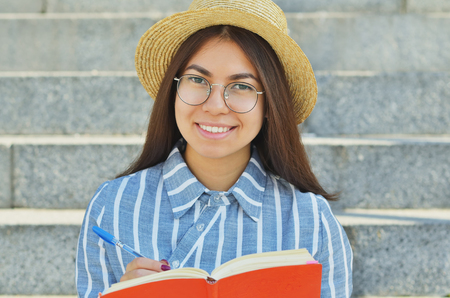 Portrait of a young Asian student in glasses with a hat dressed in a blue striped shirt who writes tasks in a notebook, and smiles at the cameraの写真素材