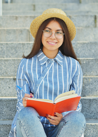 Portrait of a young Asian student in glasses with a hat dressed in a blue striped shirt that writes tasks and looks into the distance.Vertical photographyの写真素材