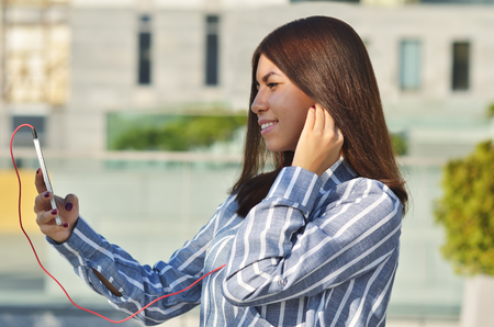 A young girl student of Asian appearance dressed in a striped shirt takes a selfie and listens to music while on the streetの写真素材