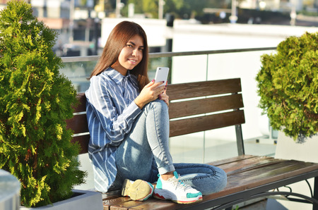A modern girl in a beautiful shirt and sneakers uses the phone and sits on a bench in the city Parkの写真素材