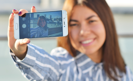 A beautiful girl in a striped blue shirt takes a selfie in the city while walkingの写真素材