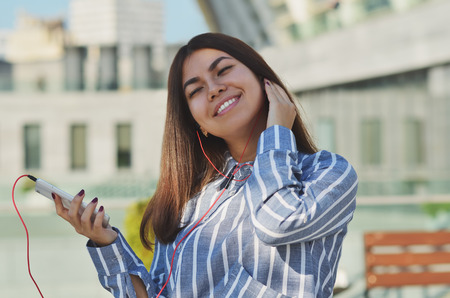 Portrait of a cheerful girl who walks around the city and listens to music from a mobile phone in vacuum headphones, raising her head up and closing her eyesの写真素材