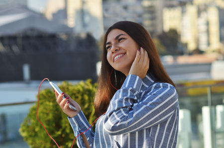 Portrait of a cheerful girl who walks around the city and listening to music from a mobile phone in vacuum headphonesの写真素材