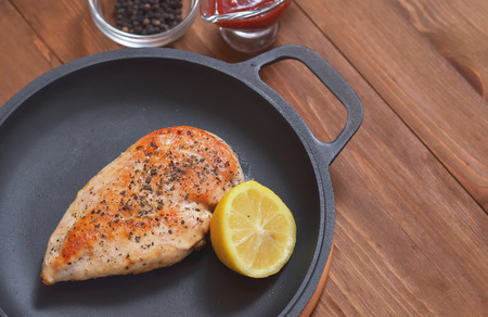 Fried chicken fillet is in a frying pan with lemon, sauce and spices on a wooden table.の写真素材