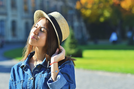 Asian girl in a hat wearing a denim jacket enjoys the sun and holds a hat with one handの写真素材