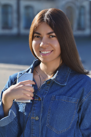 Portrait of a young girl that listens to music, Asian appearance dressed in a denim jacket with a phone in her handsの写真素材