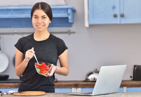 A young girl in a black t-shirt makes a fit breakfast and stirs red pepper in the bowl in a kitchen. Laptop on background. Horizontal photoの写真素材