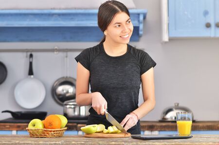 A young girl in a black t-shirt makes a fruit breakfast and cuts apple on a light wooden board in a kitchen. Orange juice and fruit in bowl is standing near. Horizontal photoの写真素材