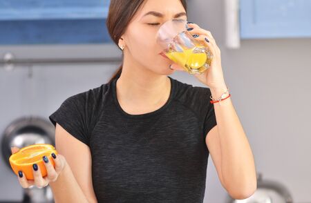 Sport Asian woman in a black t-shirt makes and drinks orange juice in the kitchen. Horizontal photoの写真素材
