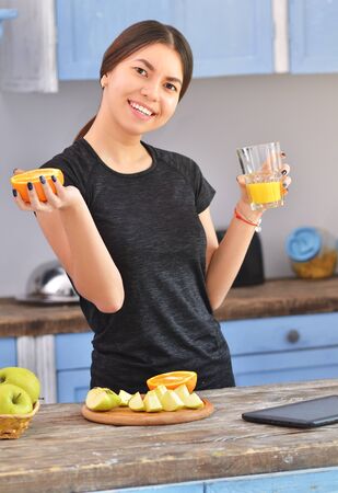 A young woman in a black t-shirt makes fit breakfast and drinks orange juice . Fruit in a bowl stands nearby. Horizontal photoの写真素材