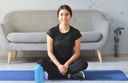 Fitness at home concept. Smiling young woman is sitting on mat with sports equipment at homeの写真素材