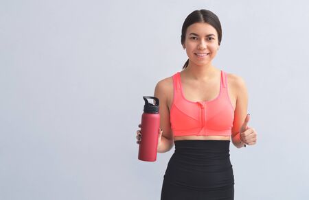 Young athletic woman in fitness equipment having a bottle of water in hand, on gray backgroundの写真素材